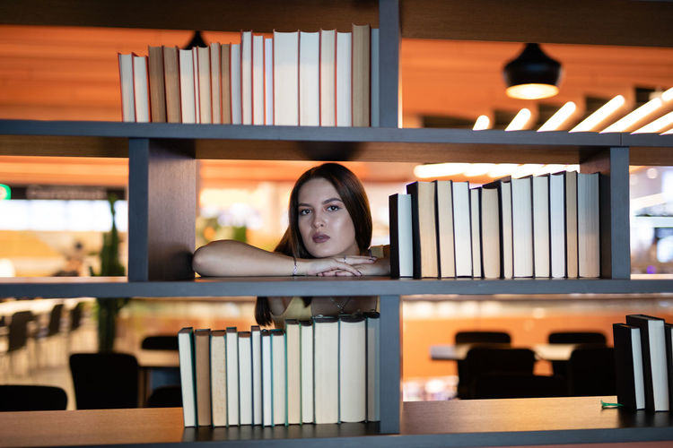 Portrait of young woman leaning on bookshelf at library