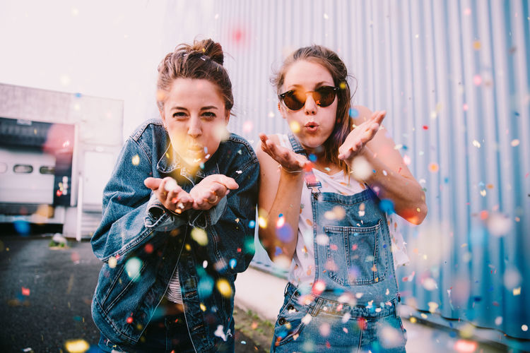 Portrait of young women blowing confetti while standing outdoors