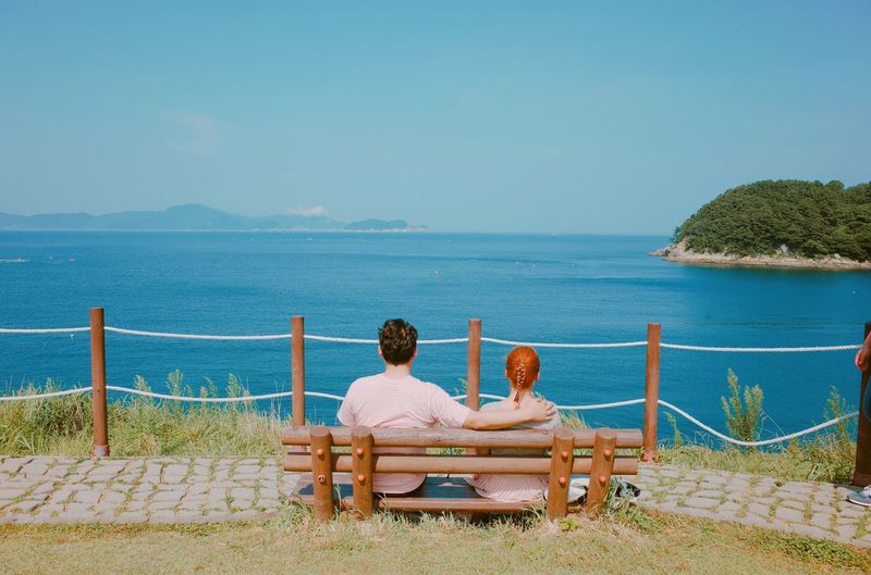 Rear view of couple sitting on bench while looking at sea against sky