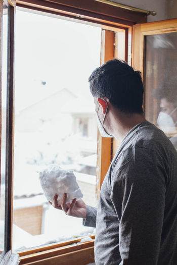 Young man with mask in quarantine with a snowballs playing through the window of his house