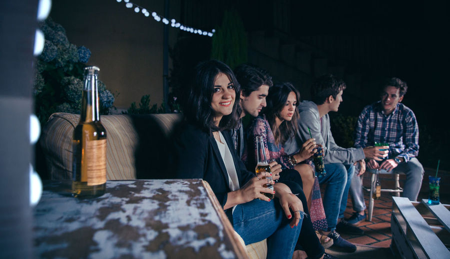 Portrait of woman smiling while having beer with friends at patio during night