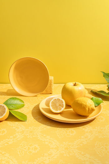 Fresh whole and halved sour lemons near apple on vivid ceramic plate on table on yellow background