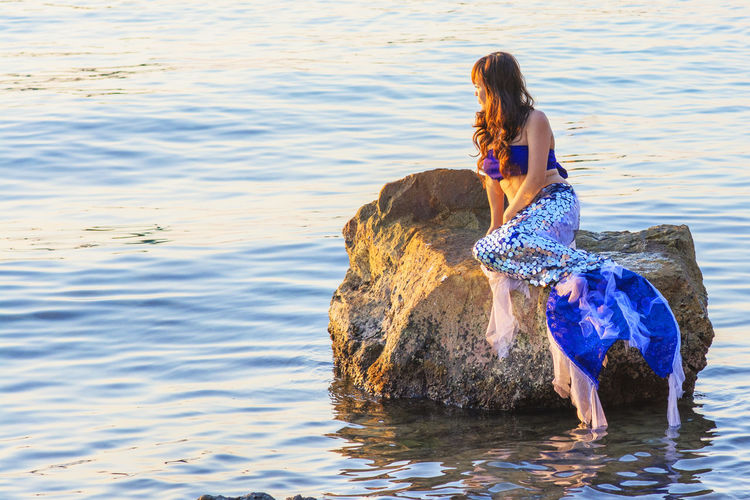 Woman in mermaid costume sitting on rock in lake