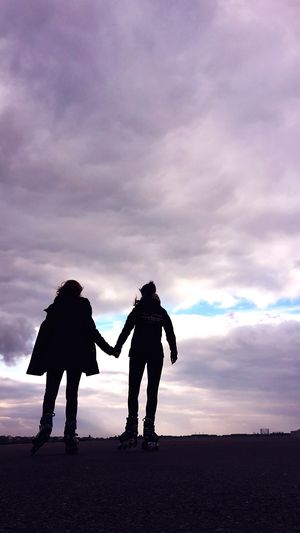 Silhouette friends holding hands while roller skating on road against cloudy sky