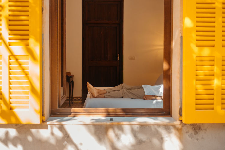 Through window of side view of unrecognizable barefooted male tourist in casual clothes lying on comfortable bed on sunny day in cozy house in mallorca