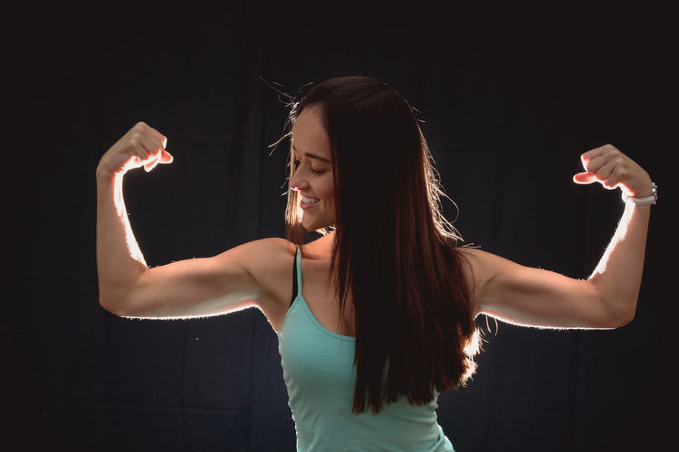 Smiling woman flexing muscles while standing against black background