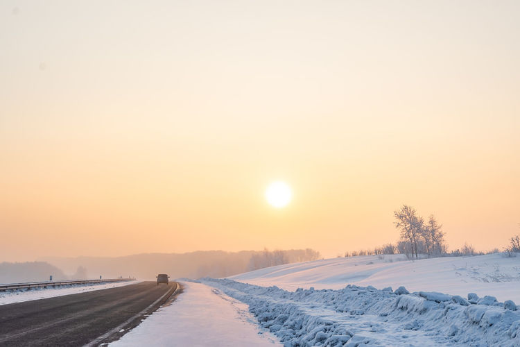 Winter frosty landscape with an asphalt road, a car walking along it and orange disk of the sun