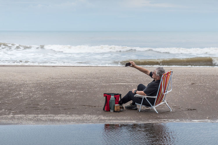 Mature man with gray hair sitting on a beach chair taking a selfie. behind waves of the sea.