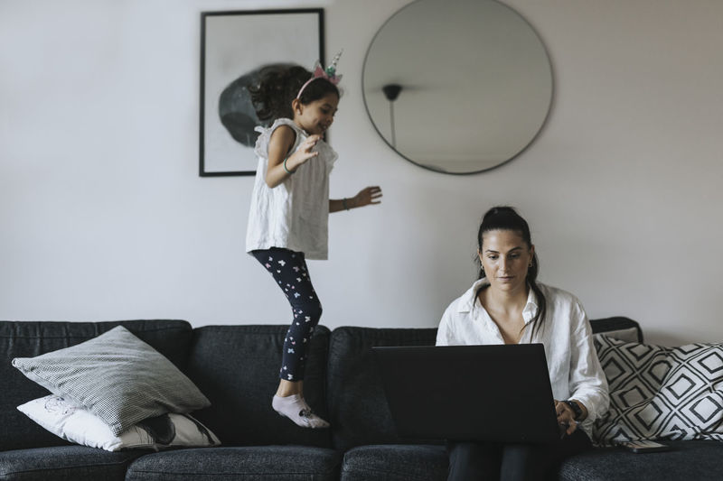Woman using laptop while daughter jumping on sofa