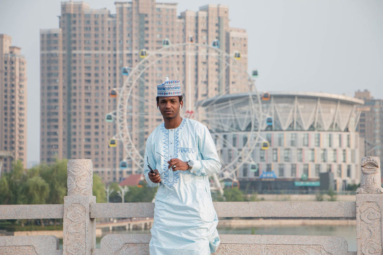 Portrait of man in traditional clothing standing against ferris wheel in city