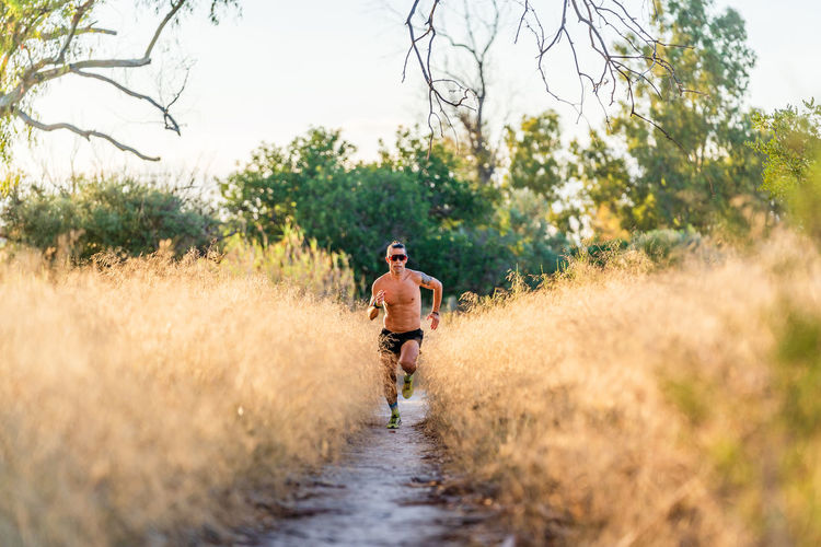 Full body shirtless male athlete with sunglasses running on narrow footpath among tall grass during outdoor workout in nature