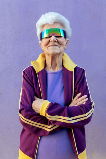 Cheerful elderly female athlete with folded arms and gray hair in sportswear and blindfold on violet background