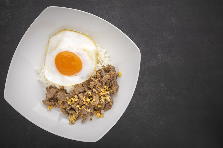 Thai food, streamed rice topped with fried beef with garlic and fried egg in ceramic plate, top view