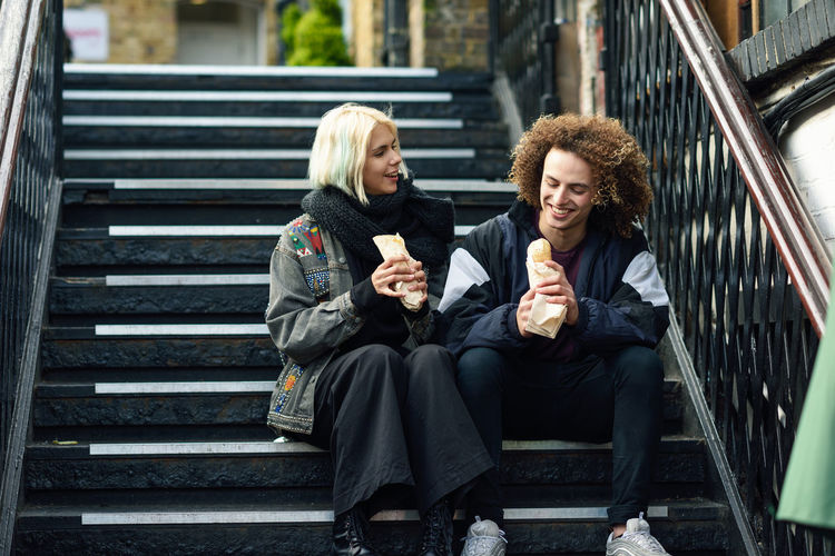 Young couple eating food while sitting on steps in city