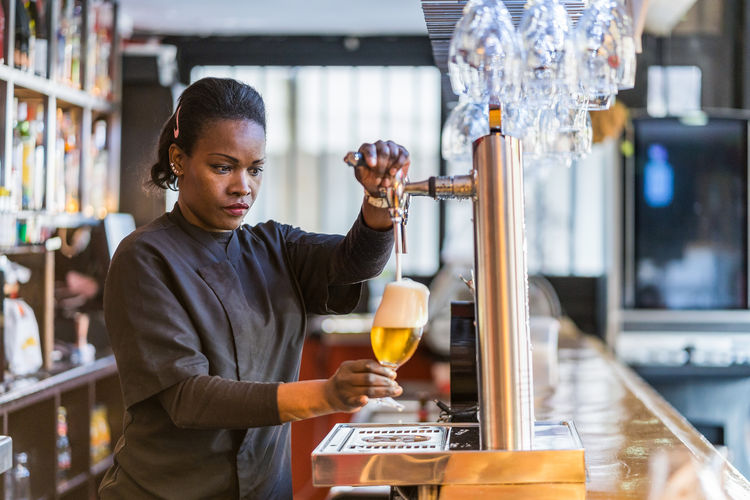 Side view of black female barkeeper pouring cold beer in glass in bar