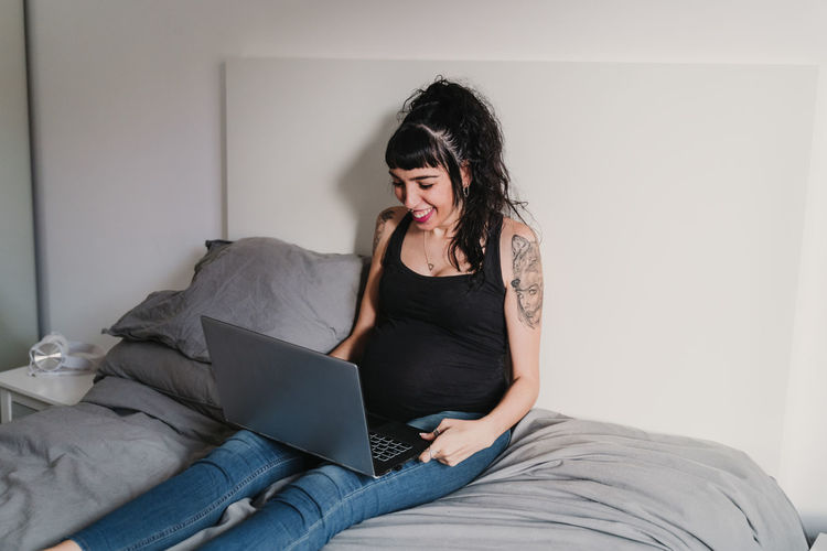 Pregnant woman using laptop while sitting on bed at home