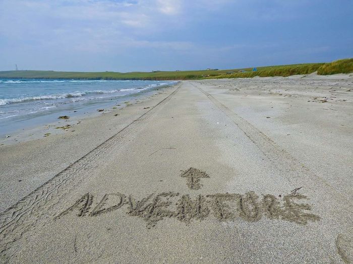 Adventure sign drawn in sand on beach against sky