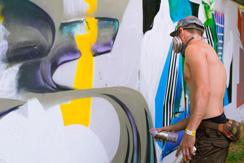 Side view of shirtless street artist painting graffiti on wall