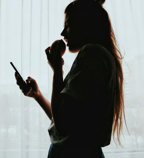 Side view of young woman using smart phone while standing against window