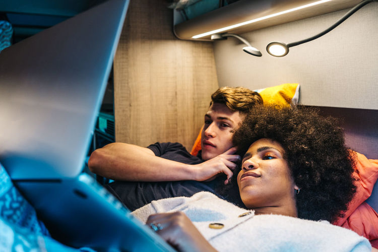 Happy romantic multiracial couple cuddling and enjoying time together while lying on bed and watching movie on laptop during travel in camper van