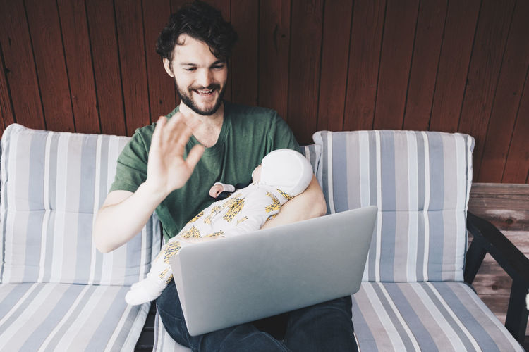 Smiling father carrying baby boy while doing video conference on laptop at holiday villa
