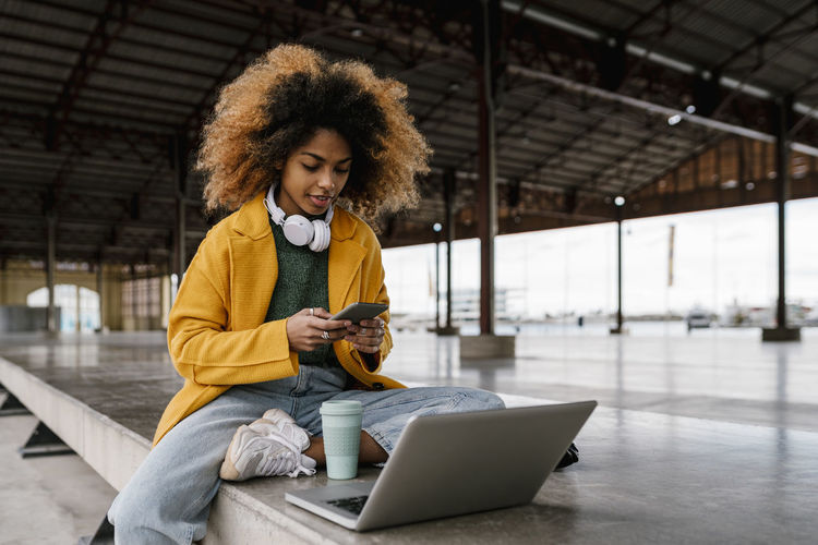 Afro woman using smart phone while sitting in front of laptop