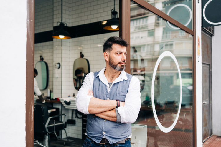 Handsome adult bearded male beauty master with arms crossed looking away standing at door of barbershop