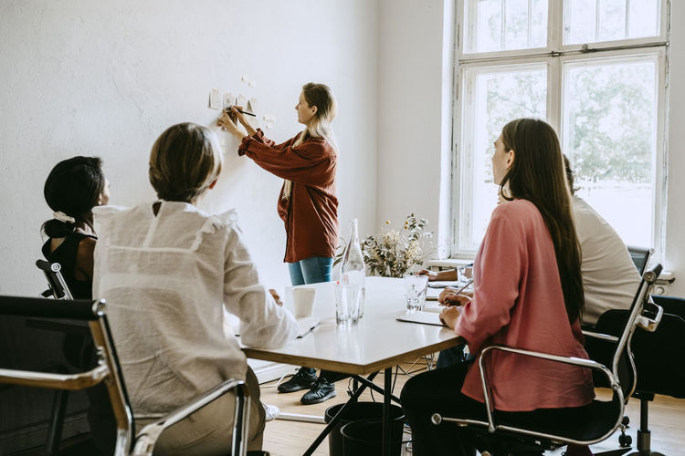 Businesswoman writing on adhesive note while explaining colleagues during seminar in office