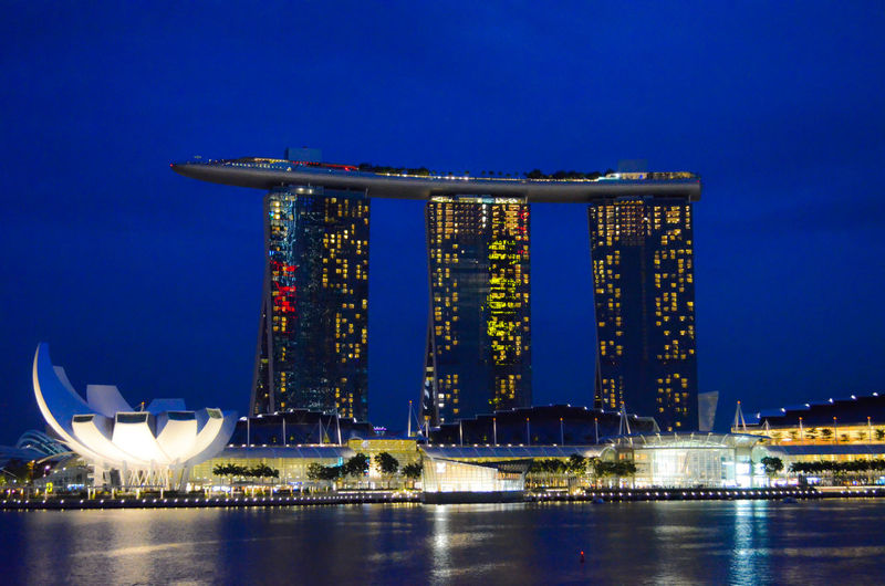 Illuminated marina bay sands in city by river against blue sky at night