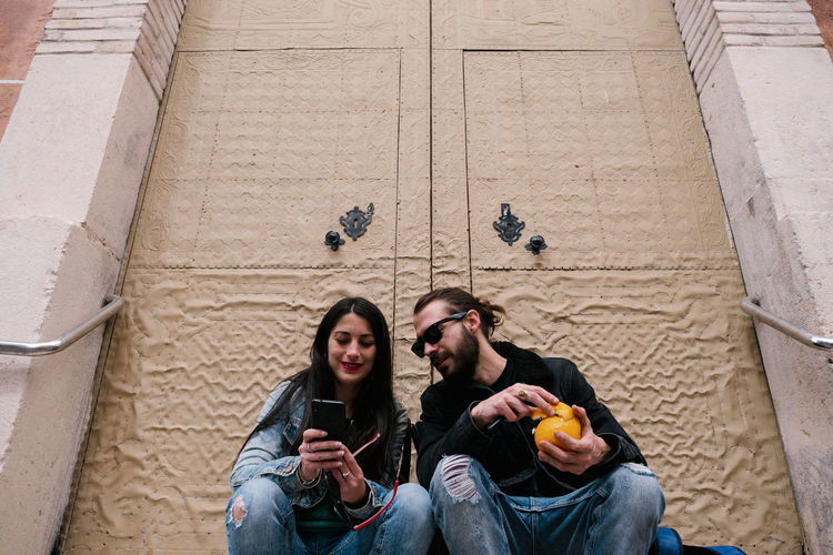 Man and woman staring at the phone eating fruit sitting in the street