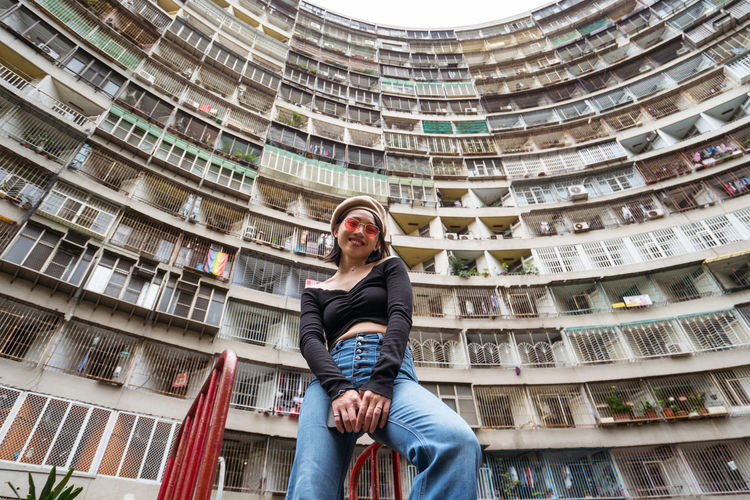 From below of young female in casual outfit standing against multistory residential building with geometric curved walls on street of kaohsiung city in taiwan