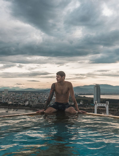 Full length of shirtless man sitting in swimming pool against sky