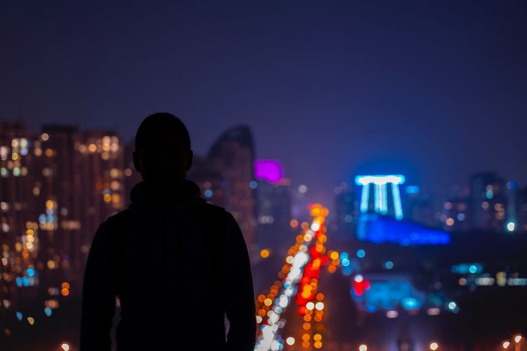 Rear view of man standing against illuminated cityscape