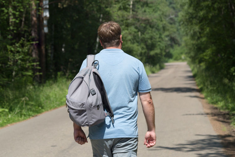 Adult male traveler with backpack is walking alone on an empty road in the middle of the forest.