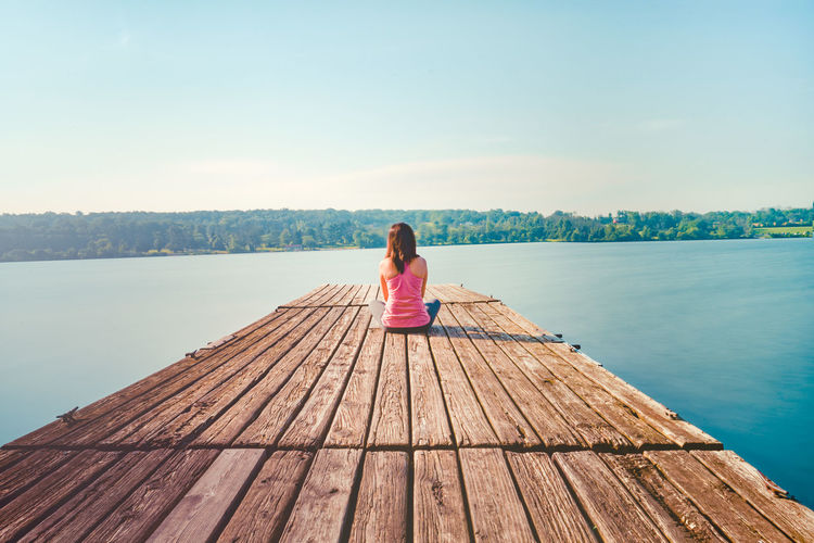 Rear view of woman sitting on pier over lake against sky
