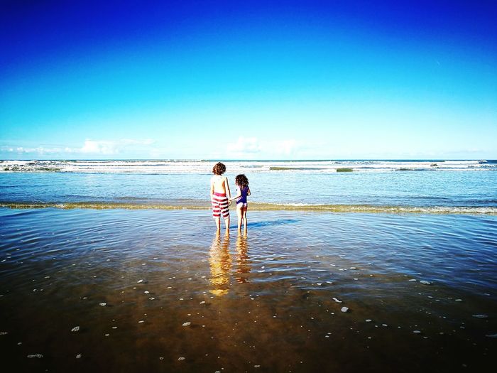 Rear view of siblings standing on shore at beach against blue sky