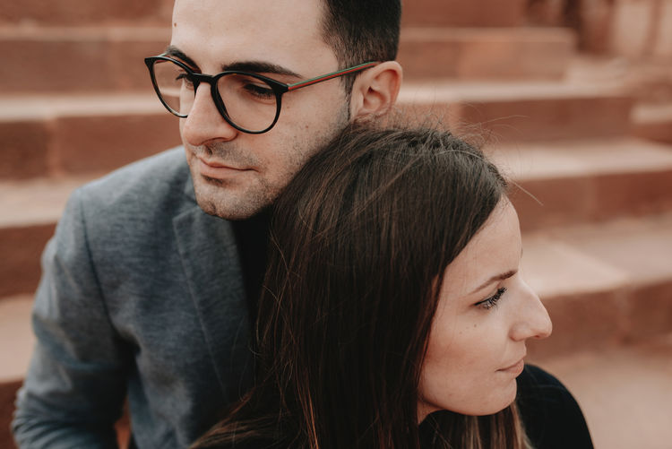From above of crop young ethnic man in elegant outfit and eyeglasses hugging calm girlfriend while resting together on stone stairs