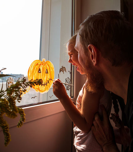 Child and father painting pumpkin on window preparing halloween little girl with dad decorating room