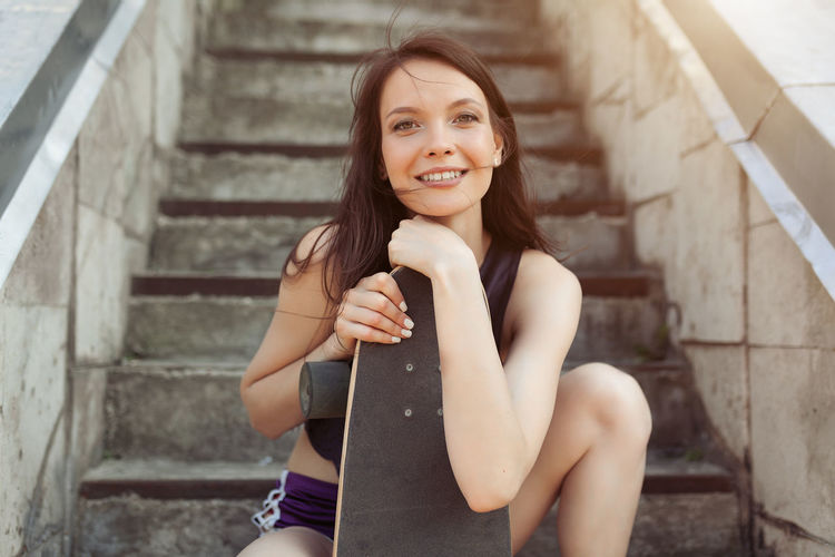 Portrait of smiling young woman with skateboard sitting by steps