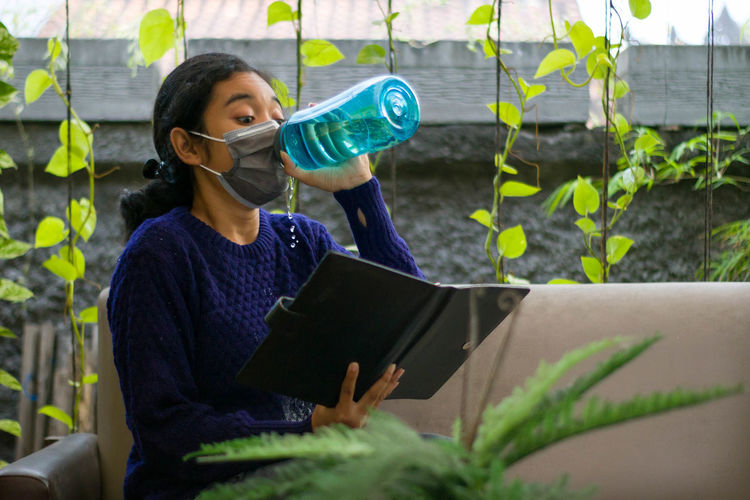 Young woman reading a book and try to drink when she still wearing a mask.