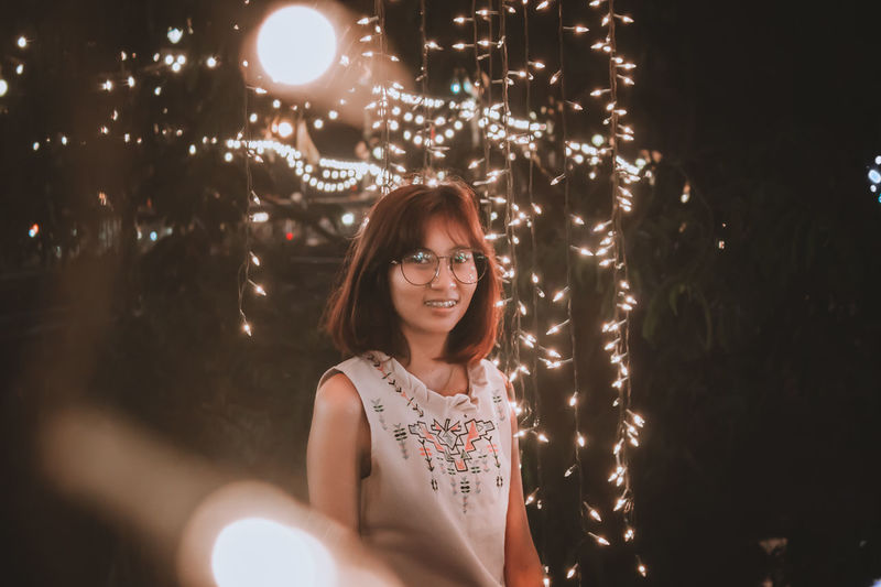 Portrait of young woman wearing eyeglasses amidst illuminated lights at night