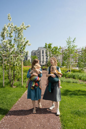 Full length of smiling caucasian mothers carrying their babies in slings at park on sunny day