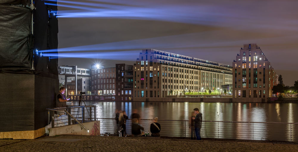 People at illuminated city by river against sky at night