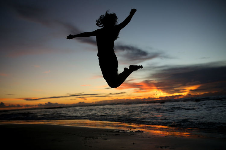 Silhouette person jumping on beach against sky during sunset