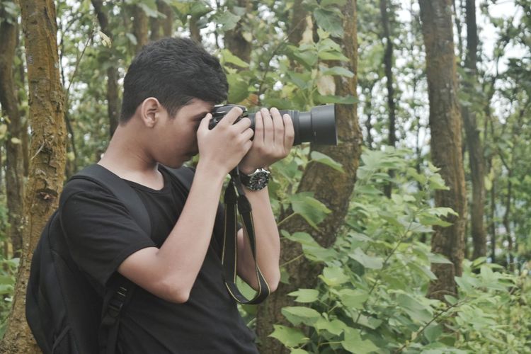 Young man photographing while standing on tree in forest