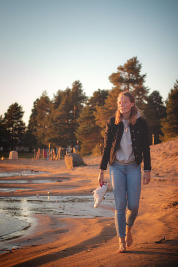 Smiling young brunette athlete walks barefoot on  sandy beach, enjoying the sunset. sotkamo, finland