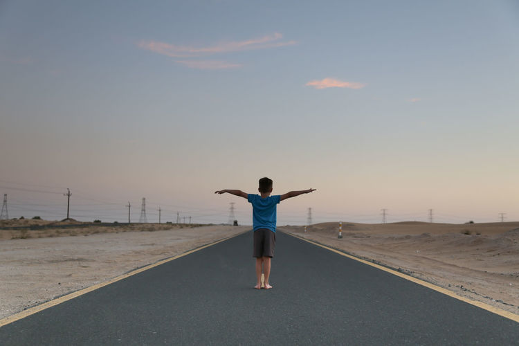 Rear view of boy with arms outstretched standing on road against sky during sunset