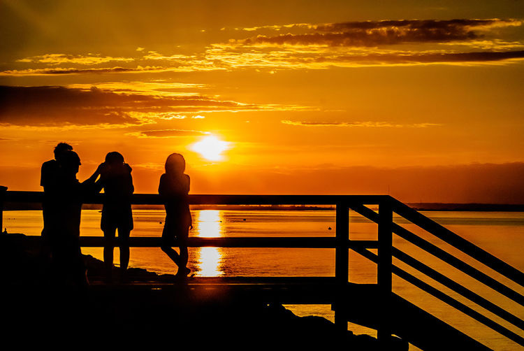 Silhouette friends standing on pier at sea shore against sky during sunset