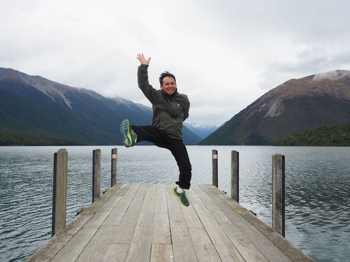 Portrait of cheerful man jumping on pier over lake against mountains