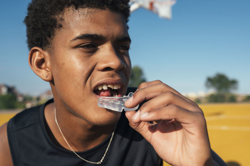 Young african american sportsman with split tooth placing transparent mouthguard in mouth in bright day on playground